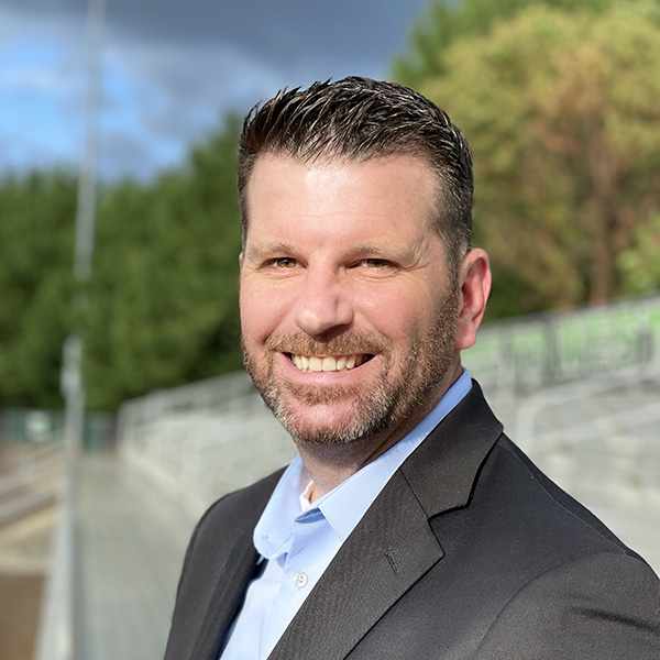 Derik Milton in a black suit and blue collared shirt outside smiling